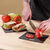 A person is slicing a tomato on a small cutting board, with sandwiches and whole tomatoes in the background. Produktbild-Vorschau