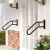 Two black U-shaped handrails mounted on a white wall next to a white door, with brickwork and greenery visible. Voir les images du produit