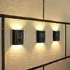Three black solar wall lights are mounted on a textured, light-colored wall with dark grout lines. The lights are illuminated, casting warm yellow light upwards and downwards. Produktbild-Vorschau