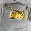 A close-up of a plush toy's chest area, showing a grey fuzzy hoodie with a gold chain and the word 'DIKKA' in gold lettering. Produktbild-Vorschau