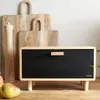 A wooden bread box with a black front is displayed on a wooden surface, with cutting boards and pears in the background. Náhled obrázku produktu