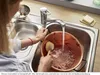 A person is washing a copper pot in a double sink with a chrome faucet running. Produktbild-Vorschau