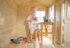 A young child stands inside a wooden playhouse next to a small table. A stuffed animal is lying on the floor. Produktbild-Vorschau