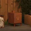A corten steel garden stove is shown on a patio next to a stack of firewood and a wicker furniture piece. Náhľad obrázku produktu