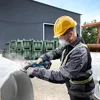 A construction worker is using an angle grinder on a large white object. The worker is wearing safety gear including a hard hat, safety glasses, and a dust mask. The background shows a large industrial building and some green metal structures. Náhľad obrázku produktu