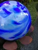 A close-up of a decorative glass sphere with blue and white swirls, resting on a metal base with petal-like feet, set in an outdoor garden environment with greenery in the background. Anteprima dell’immagine prodotto