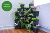 A modular vertical planter system filled with various plants and flowers, placed in a corner against a wall. Produktbild-Vorschau
