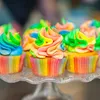 A close-up view of several rainbow-frosted cupcakes on a clear glass cake stand. Náhled obrázku produktu