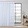 A white sheer curtain hangs in front of a large window with a black frame. A small wooden table with decorative items is placed to the right of the window. Produktbild-Vorschau