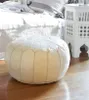 A cream-colored leather pouf is placed on a wooden floor in front of a bed. A white side table is visible to the right of the pouf. Anteprima dell’immagine prodotto