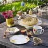 An Easter-themed table setting with decorative eggs, flowers, baked goods, and ceramic figurines. Náhled obrázku produktu