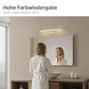 A woman in a bathrobe stands in front of a mirror in a bathroom, with a light fixture above the mirror. Produktbild-Vorschau