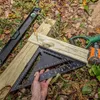 A person's hand holding a black speed square next to wooden planks and construction tools on the ground. Produktbild-Vorschau
