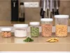 Several clear plastic food storage containers with white lids are arranged on a countertop. The containers hold various food items such as spices, flour, dried herbs, chickpeas, and pasta shells. A small pile of dried herbs and a few crackers are placed in Náhľad obrázku produktu