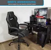 A black gaming chair in front of a computer desk with a monitor, speakers, and PC tower. Produktbild-Vorschau