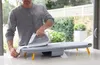 A man is ironing clothes on a portable ironing board on a table, with a window and greenery in the background. Náhľad obrázku produktu