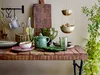 A rustic wooden table laden with various kitchen items including a green juicer, pitcher, glasses, and bowls. Produktbild-Vorschau