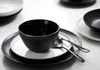A close-up overhead view of a black textured bowl placed on a white plate, which is on a black textured charger plate. Two spoons are resting on the white plate. Another white plate with a black bowl is visible in the background. Náhled obrázku produktu