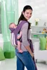 A woman is carrying a baby in a purple baby carrier in a room. Produktbild-Vorschau