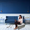 A woman is sitting on a folded mattress on the floor, using a laptop. The scene is set in a modern living room with a blue sofa and blue walls. Náhľad obrázku produktu