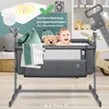 A baby bassinet with a baby, teddy bear, and pillows inside, set against a backdrop of nursery items and a wooden toy. Produktbild-Vorschau
