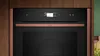 A close-up view of a modern oven with a black front panel and copper accents, framed by dark wood. Produktbild-Vorschau