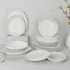 A collection of white ceramic plates and bowls are neatly arranged on a light gray surface against a white textured background. Voir les images du produit