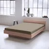 A bed with taupe fitted sheet in a minimalist room with large windows. Produktbild-Vorschau