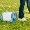 A rolled-up picnic blanket is placed on a grassy field with small white flowers. A person's legs are visible in the background, wearing dark pants and white sneakers. Náhľad obrázku produktu