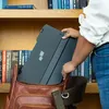 A person is placing a black portable monitor into a brown leather bag, with bookshelves in the background. Voir les images du produit