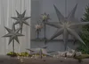 A collection of grey paper star lanterns of various sizes are displayed on a wooden surface, some on stands, with fairy lights and gift boxes. Podgląd zdjęcia produktu