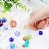 A hand is picking up a glass marble from a collection of marbles on a white surface, with green leaves in the background. Náhľad obrázku produktu