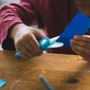 A child's hands are shown cutting blue paper with turquoise safety scissors on a wooden table. Náhled obrázku produktu
