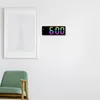 A digital clock with a colorful display is mounted on a white wall next to a framed picture of the Empire State Building. A green armchair is visible in the foreground. Náhled obrázku produktu