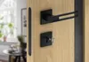 A close-up shot of a modern black door handle and lock set mounted on a light wood door. The background shows a blurred interior scene with a window and furniture. Produktbild-Vorschau