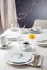 A table setting with plates, cups, and cutlery, arranged for a meal, with a vase of branches and a glass of orange juice. Produktbild-Vorschau