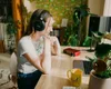 A young woman wearing headphones sits at a desk, looking at a laptop. There is audio equipment, a mug, and plants on the desk. Anteprima dell’immagine prodotto
