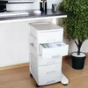 A white plastic rolling drawer unit with five drawers is shown under a desk. One drawer is partially open, revealing papers inside. A potted plant is visible to the right. Produktbild-Vorschau