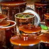 A close-up view of several brown ceramic fermentation crocks, one of which is filled with pickles and brine, with a cucumber and garlic visible. Voir les images du produit