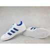 A pair of white Adidas sneakers with blue stripes and laces, shown on a light gray concrete background. Podgląd zdjęcia produktu