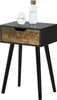 A black side table with a wooden drawer front and three black legs, shown against a white background. A glass of water and a bottle of water are on top of the table. Náhled obrázku produktu