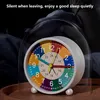 A colorful analog alarm clock with a clear dome cover sits on a wooden surface, with a dark, blurred background suggesting a bedroom. Produktbild-Vorschau