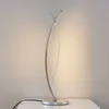 A modern, silver-colored LED table lamp with a curved design is shown on a white surface against a textured, off-white wall. Produktbild-Vorschau
