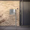 A modern silver mailbox on a stand in front of a brick wall and a dark gray door. Náhled obrázku produktu