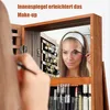 A woman is applying makeup in front of a jewelry cabinet with a mirror. Náhľad obrázku produktu