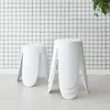 Two white stools are placed on a light gray floor in front of a white wall with a black grid pattern. A green plant hangs from the top right corner. Produktbild-Vorschau