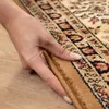 A hand is shown lifting the edge of a rug, revealing the texture and pattern. Produktbild-Vorschau