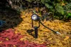 A black outdoor spotlight with a white LED light is staked into red and yellow mulch, with green foliage in the background. Anteprima dell’immagine prodotto