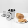 A stainless steel meat cooker with a lid and spring mechanism, next to a cooked meatloaf and two slices of meat on a white plate, all on a white surface. Produktbild-Vorschau