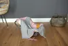 A baby car seat with a pink and gray blanket and a small pink toy on a wooden floor, with a basket and a wicker chair in the background. Produktbild-Vorschau